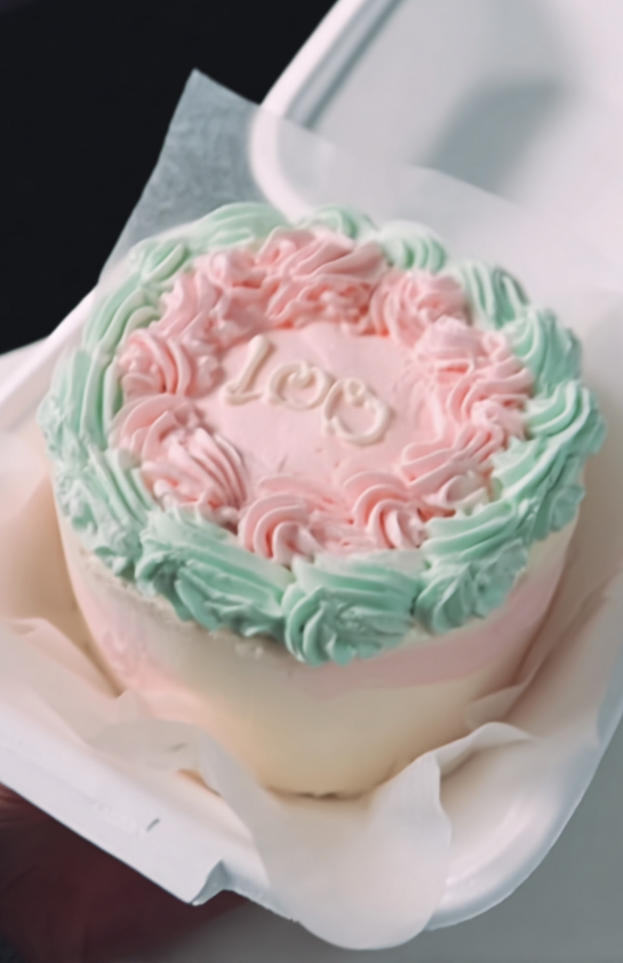 Un bento cake pastel avec bordures de crème rose et verte et un message “100” écrit en blanc, parfait pour célébrer.