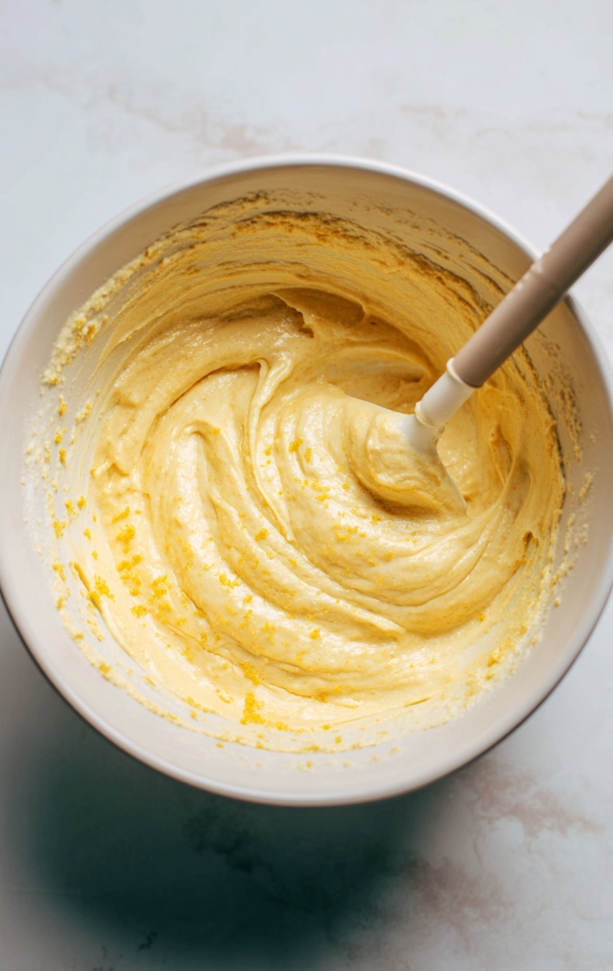 Saladier contenant la pâte à madeleines avec spatule, on voit la texture lisse et quelques zestes de citron.