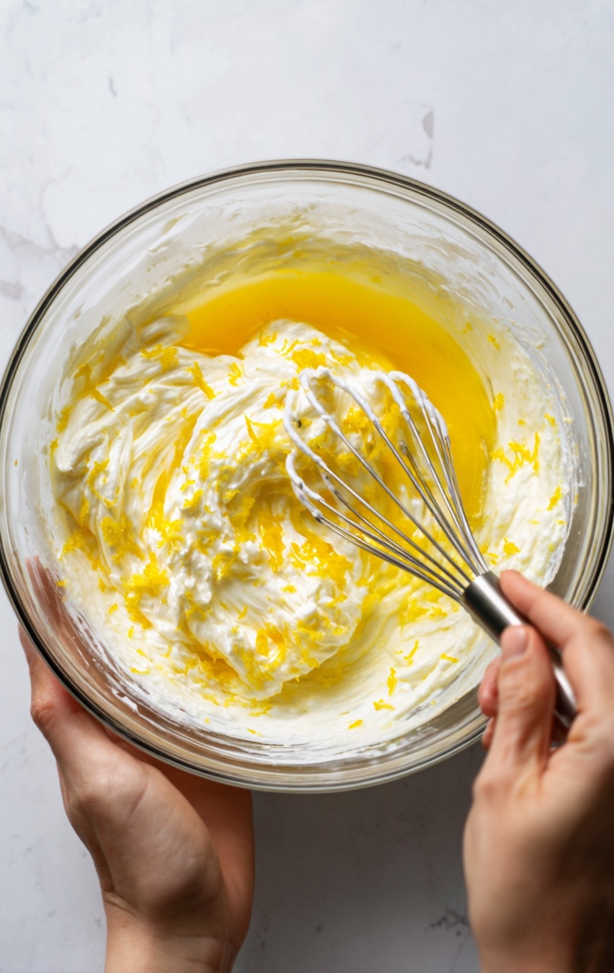 Vue de dessus : un saladier avec une préparation blanche et jaune (blancs montés et éléments citronnés) et une main tenant un fouet.