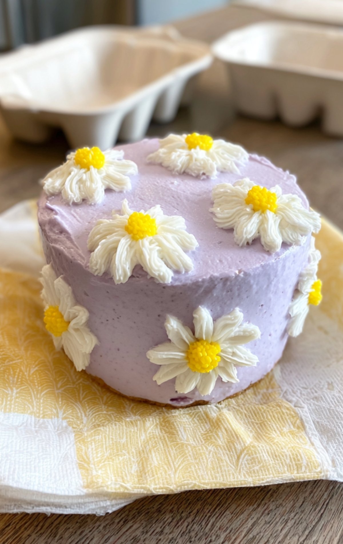 Gâteau glacé lavande orné de fleurs pochées blanches au cœur jaune, étape décoration du bento cake.