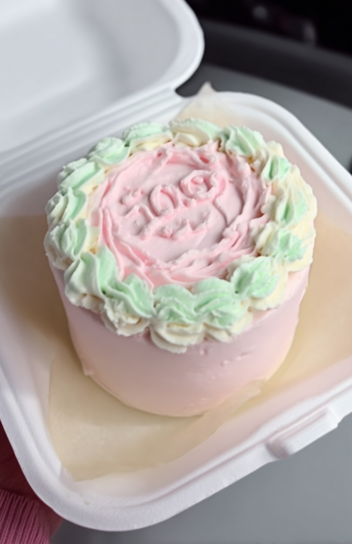 Un bento cake rose décoré de crème pastel verte et blanche dans une boîte à emporter, prêt à offrir ou à dévorer.
