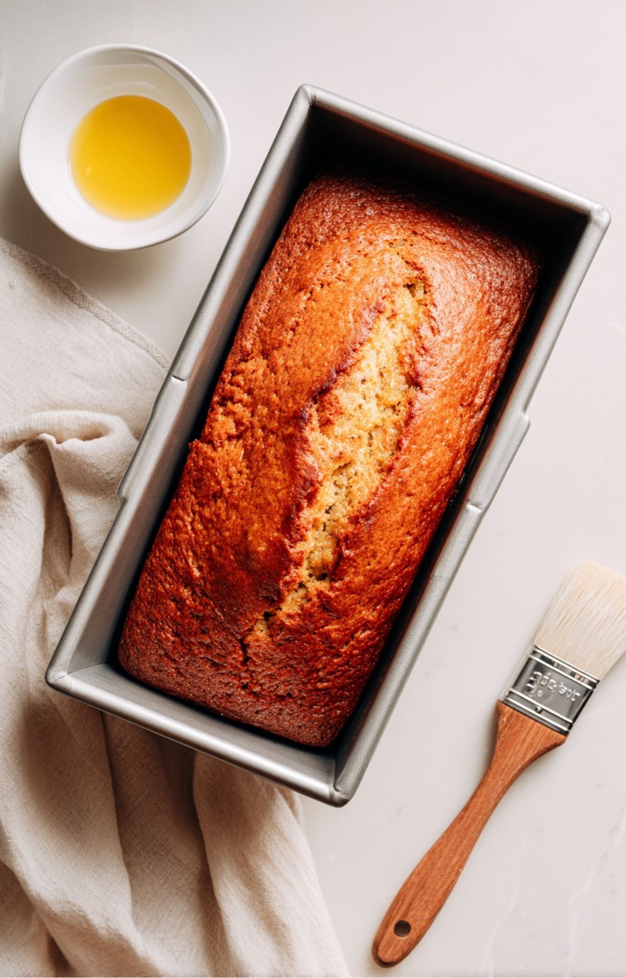 Cake doré juste sorti du four, posé dans un moule à cake avec un pinceau de cuisine et un bol d’ingrédient à côté