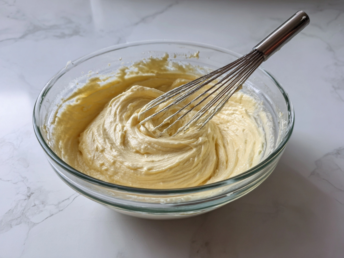 Préparation de la pâte dans un bol en verre, où l'on fouette les œufs et le sucre avec un fouet en inox pour obtenir un mélange homogène.