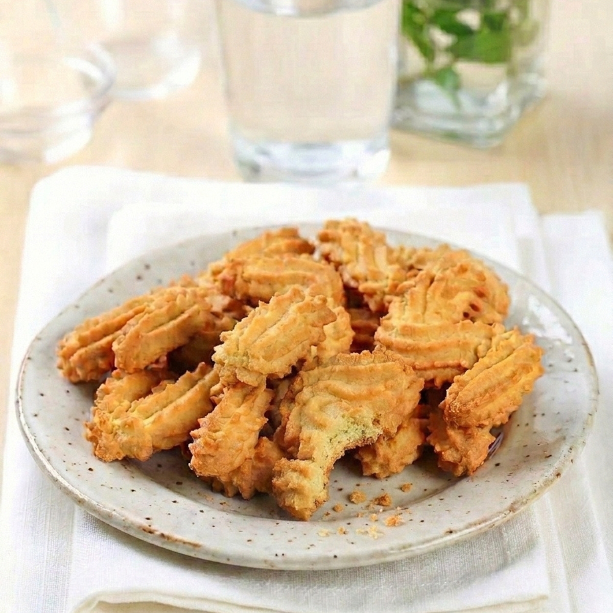 Une assiette de gateau sec spritz dorés posée sur un tissu blanc à côté d'un verre d'eau frais.
