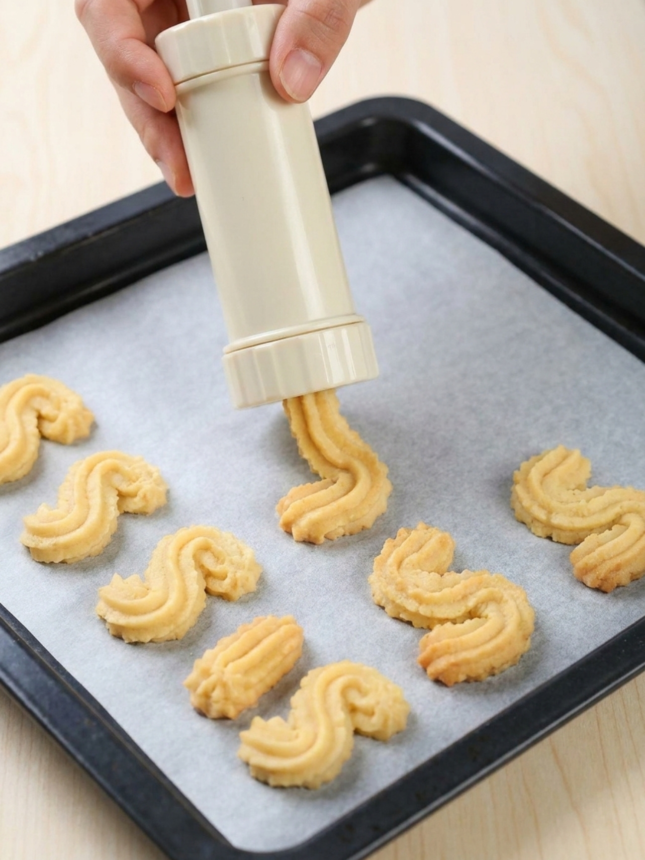 Une main qui utilise une presse à biscuits pour pocher un gateau sec spritz en forme de S sur une plaque.