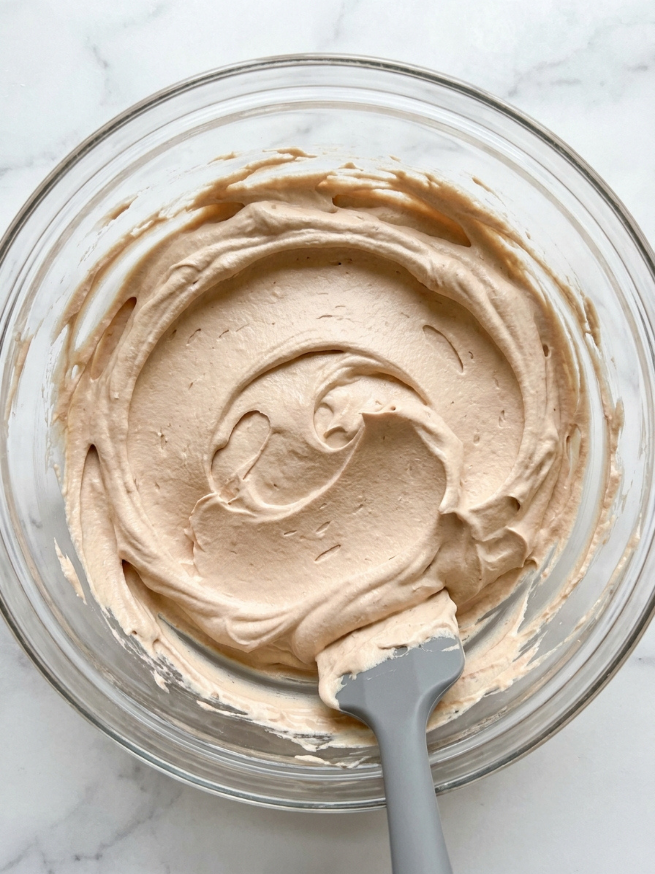 Une crème fouettée couleur beige clair dans un bol en verre mélangée avec une spatule grise.