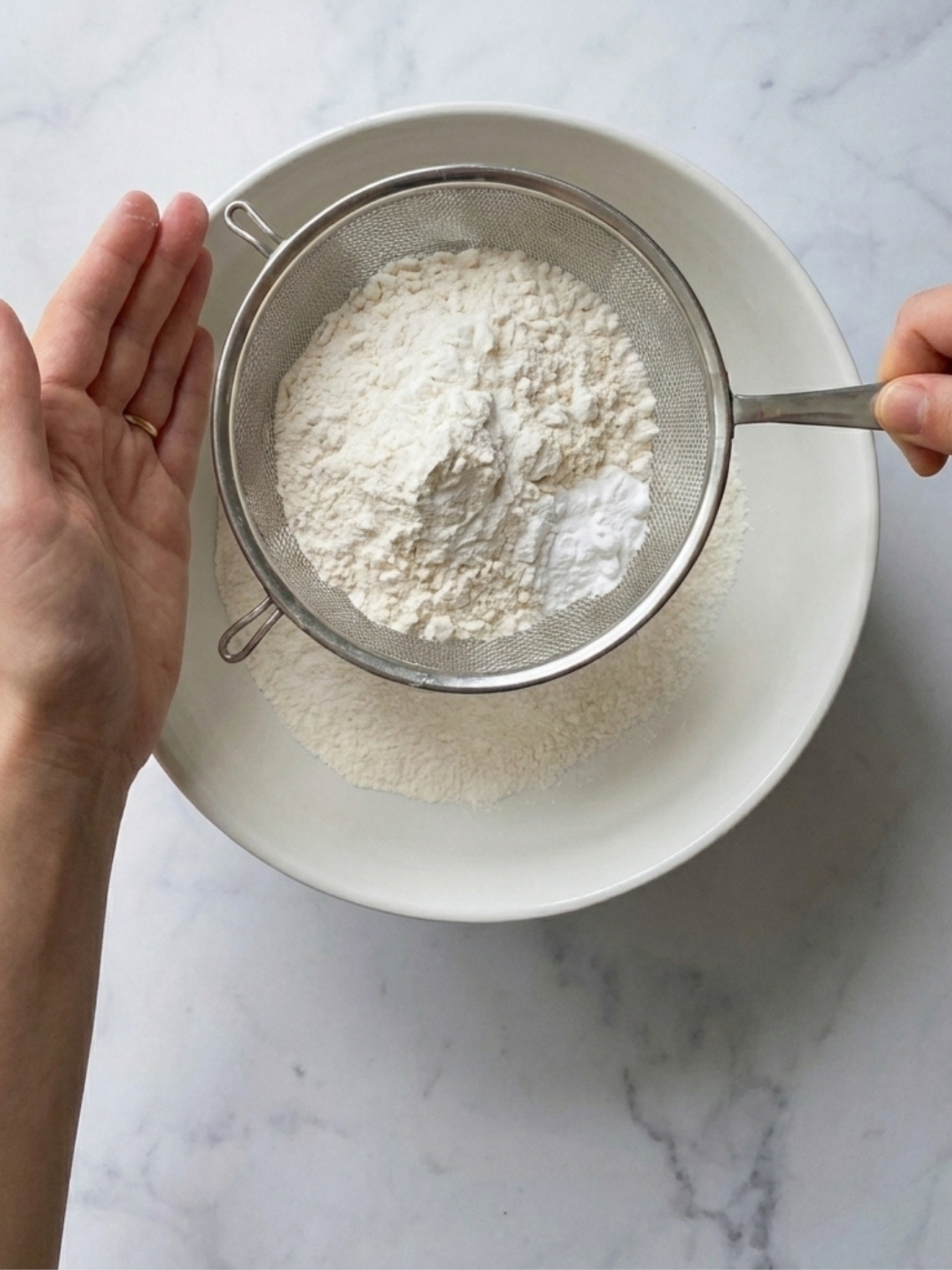 Mains qui tamisent de la farine blanche au-dessus d'un bol pour un gateau yaourt banane.