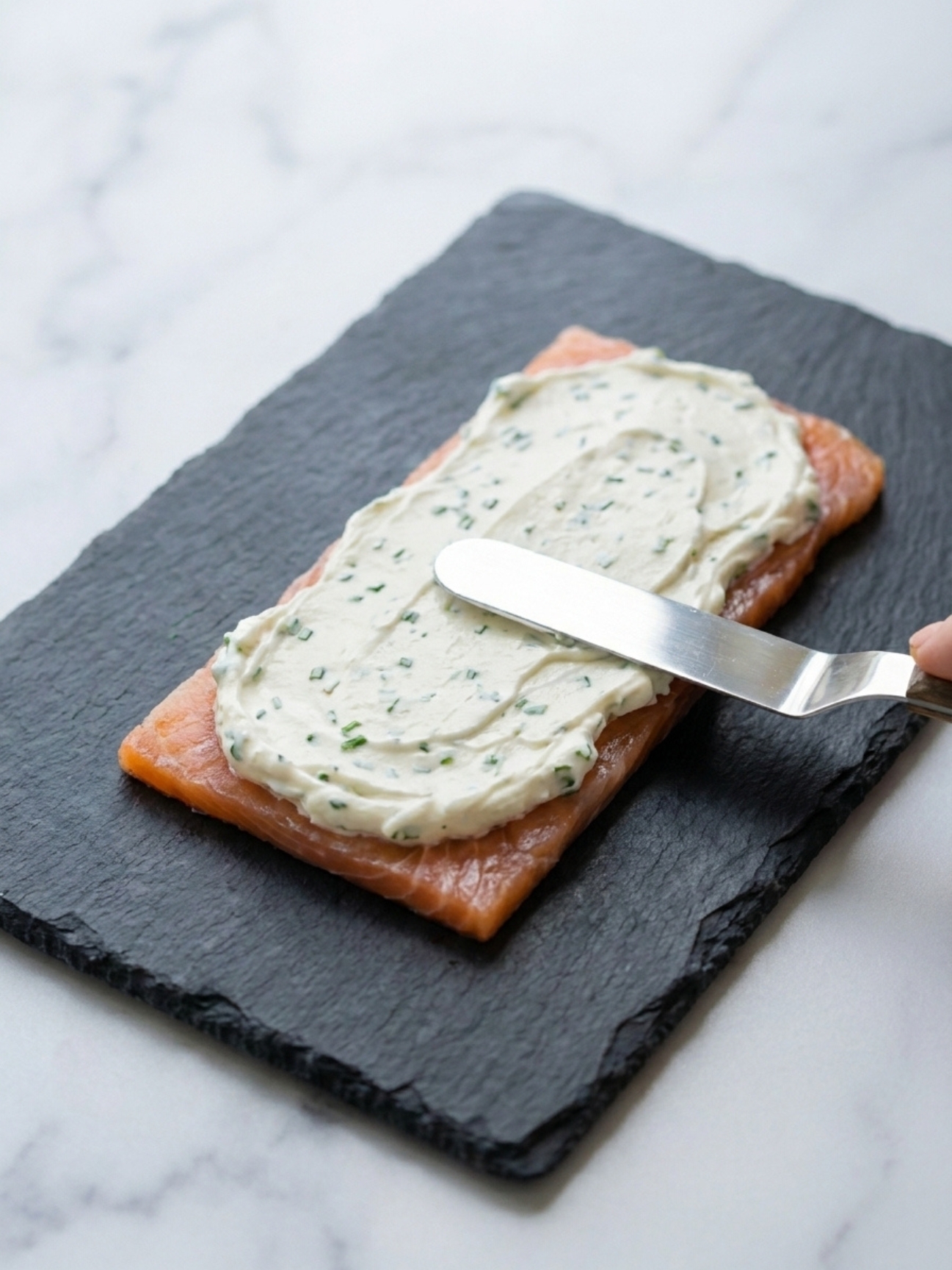 Étalage d'une crème blanche aux herbes avec une spatule sur une tranche de saumon fumé posée sur une ardoise noire pour un mille feuille saumon.