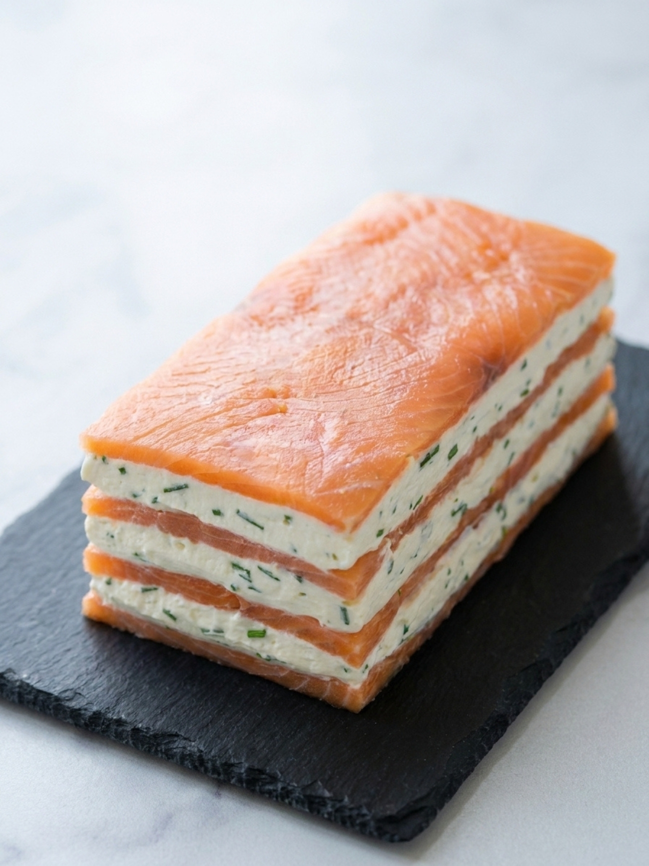 Un bloc de mille feuille saumon prêt à être découpé, montrant des couches alternées de saumon et de crème sur une ardoise.