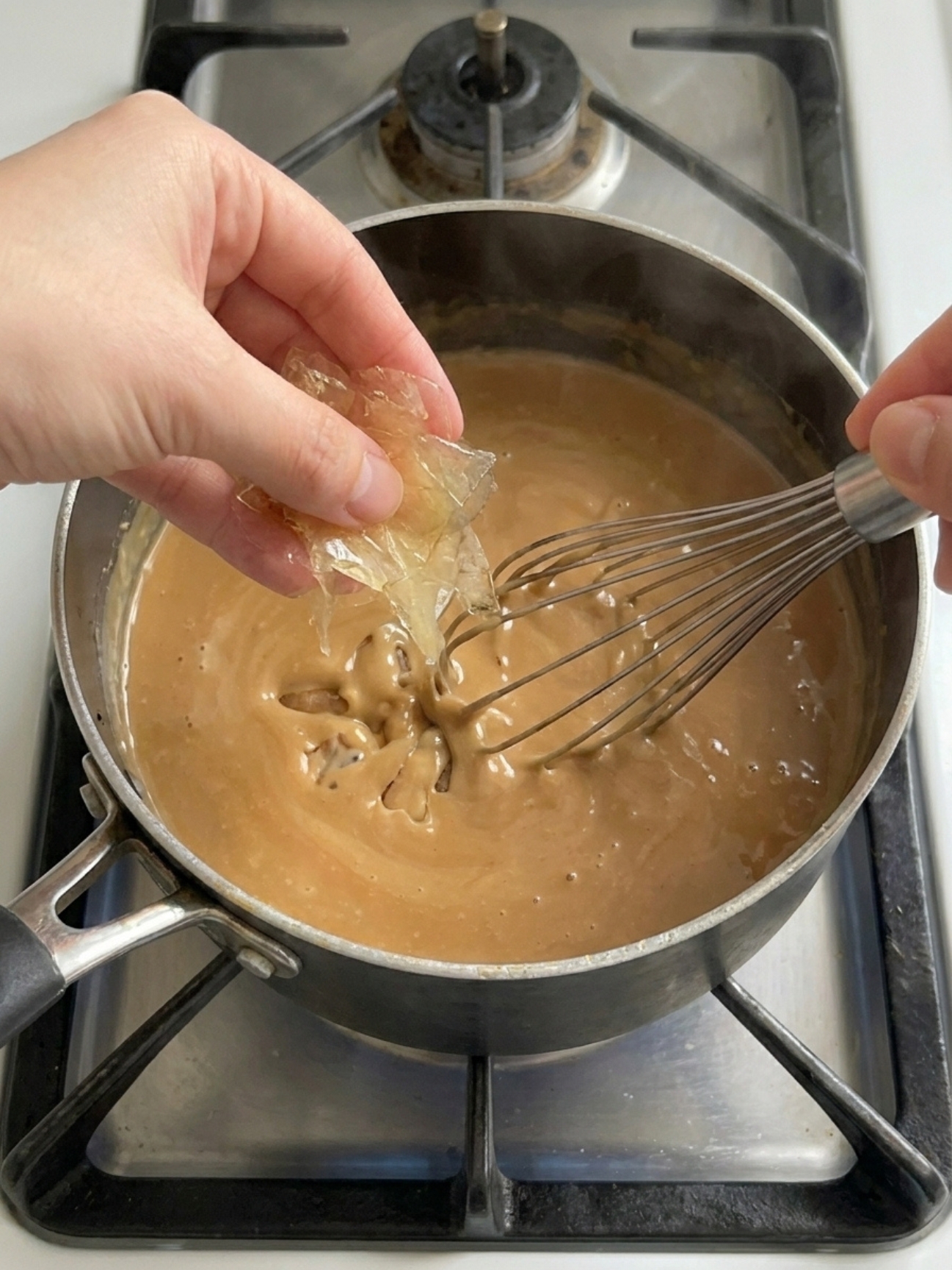 Une main ajoute des feuilles de gélatine essorées dans une crème au caramel, recette avec gélatine en feuille.