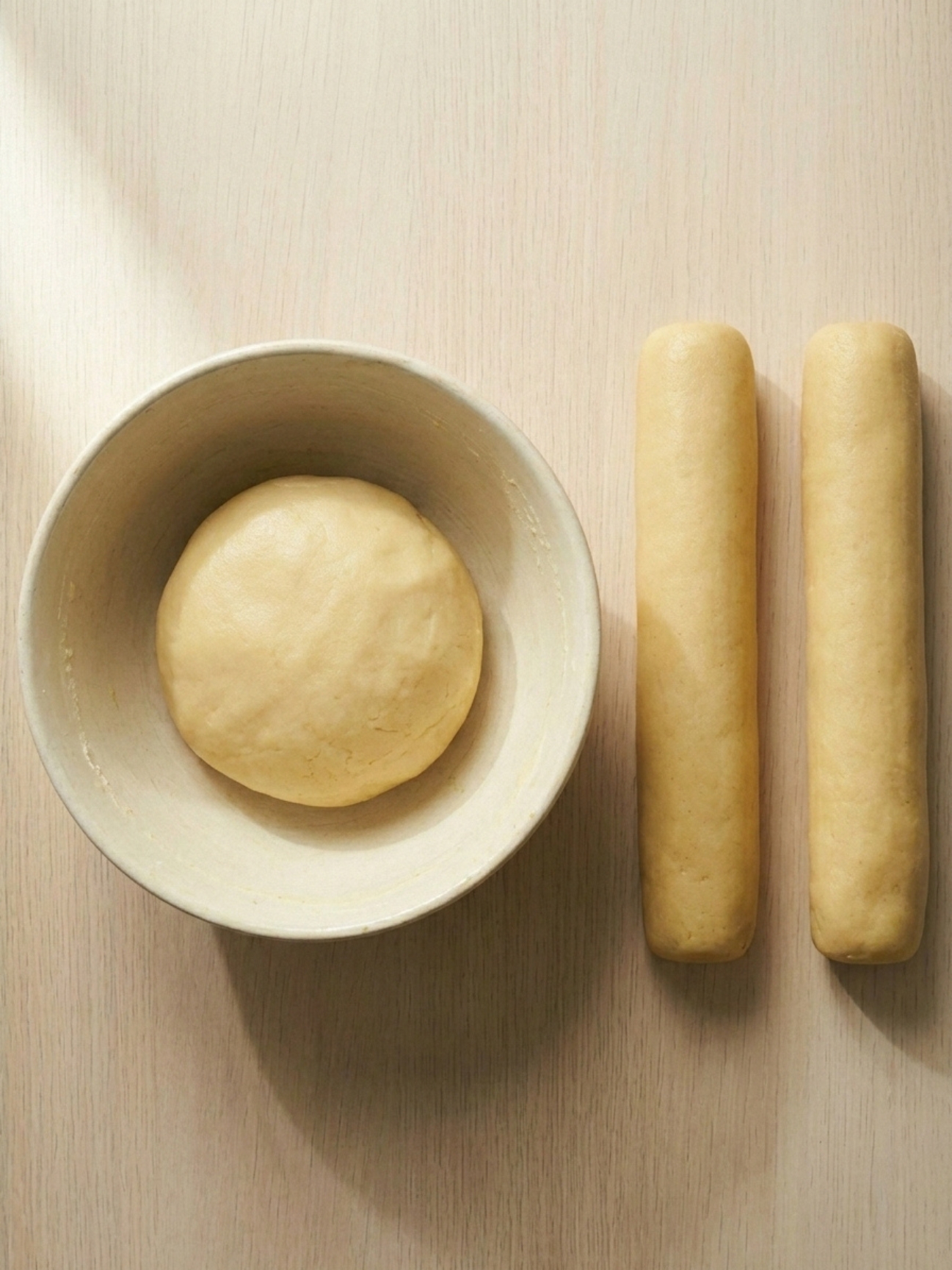 Un bol blanc avec une boule de pâte lisse à côté de deux boudins de pâte façonnés sur la table.
