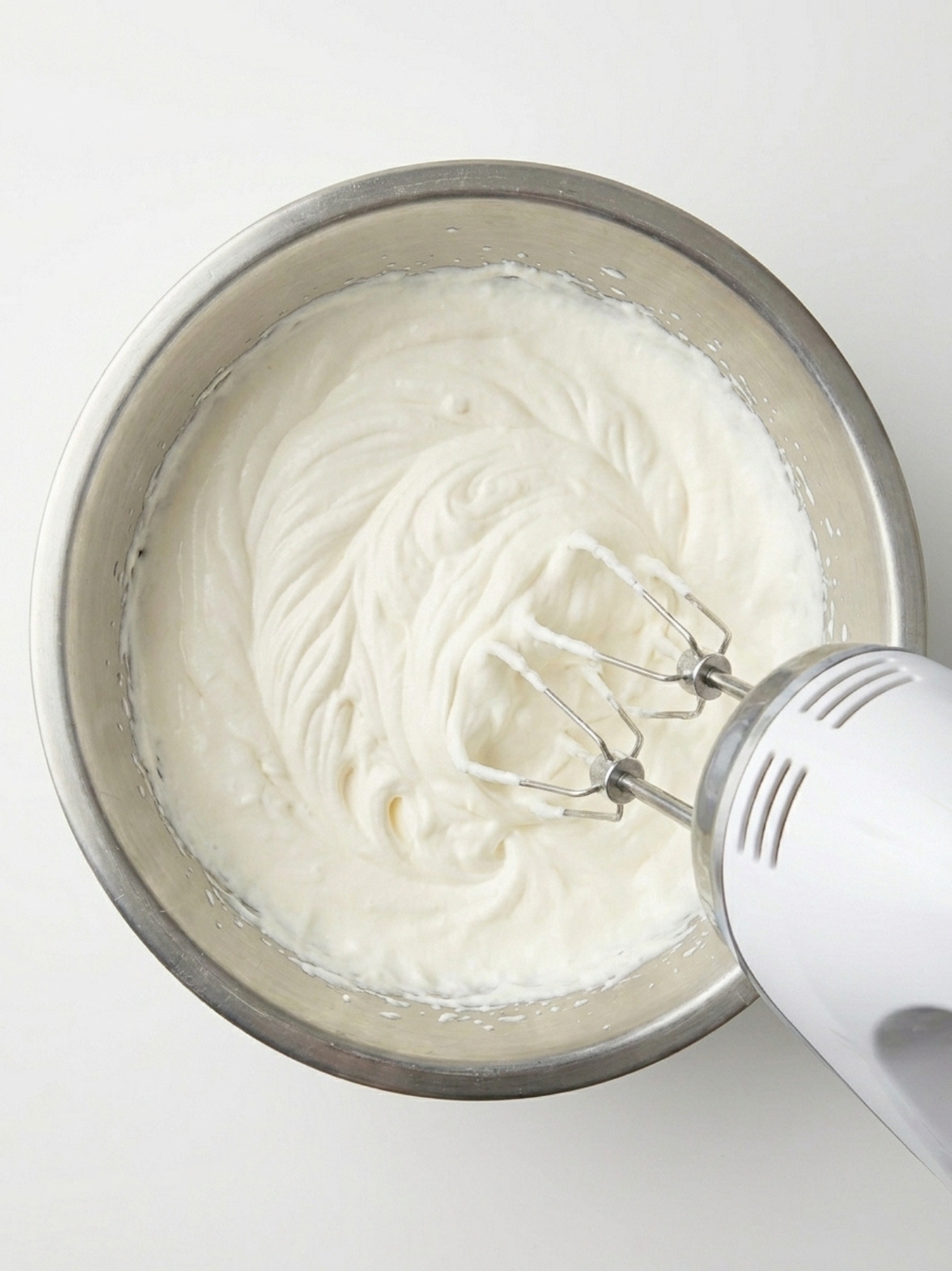 Un batteur électrique blanc fouettant une crème mascarpone blanche et ferme dans un bol.