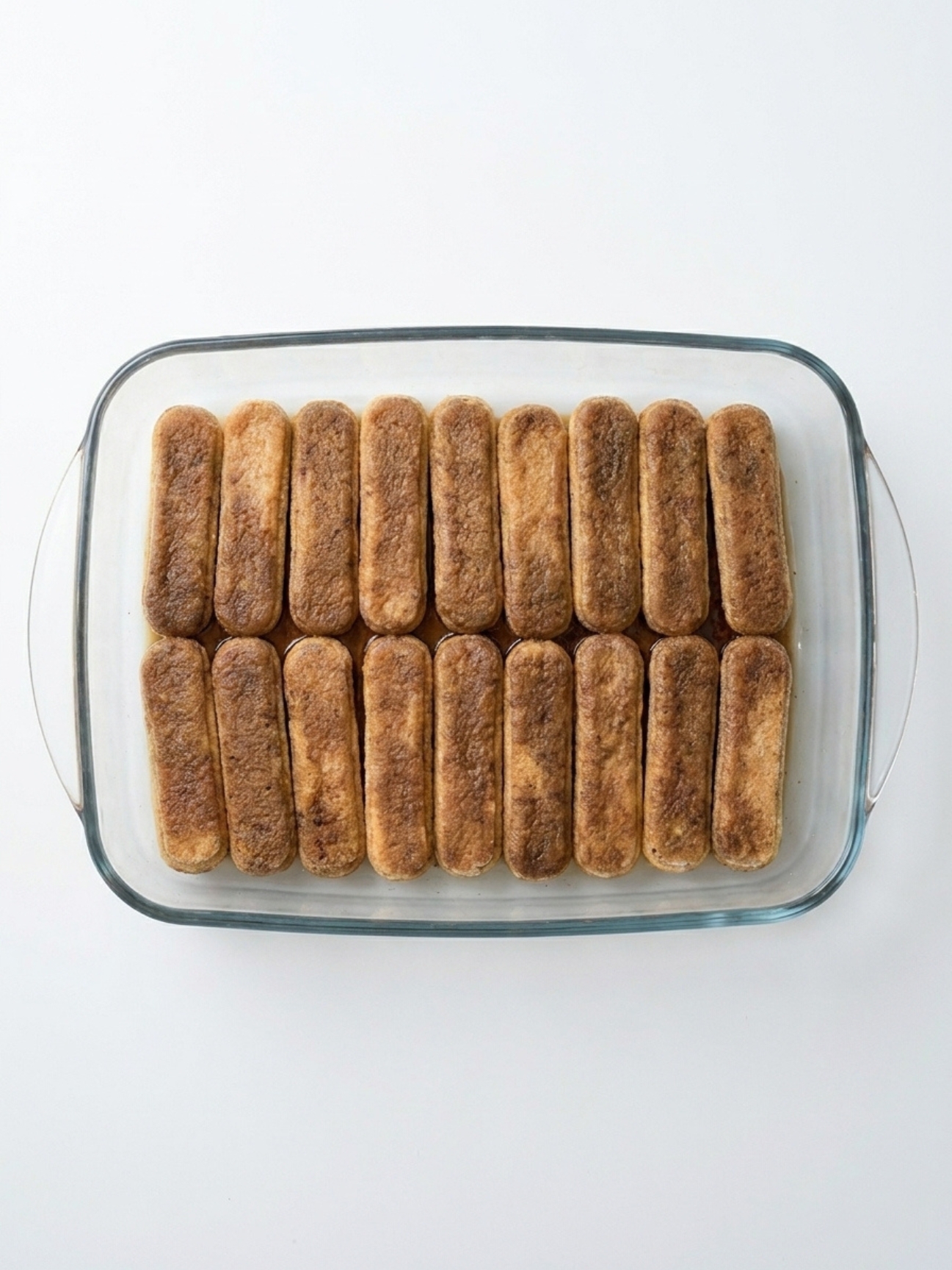 Biscuits à la cuillère imbibés de café alignés au fond d'un plat transparent pour un tiramisu.