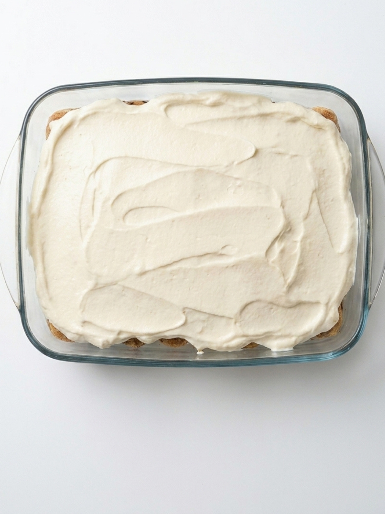 Une spatule étale une couche de crème blanche sur des biscuits dans un plat pour un tiramisu.